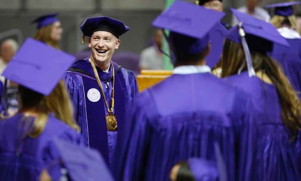 校长Victor J. Boschini, Jr.她面带微笑，身着校服，向戴着紫色帽子、身穿紫色长袍的TCU毕业生致意